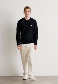 Un tânăr merge, purtând un tricou negru cu mânecă lungă cu logo-ul "BOSS", pantaloni de culoare crem și adidași albi, stând în fața unui perete alb simplu.