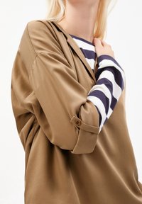 Donna che indossa un blazer color cammello sopra una maglietta a righe blu scuro e bianche con maniche lunghe, con le maniche del blazer arrotolate e abbottonate a gomito.
