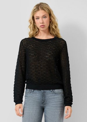 Jonge vrouw met golvend blond haar, gekleed in een zwarte gebreide trui en lichtblauwe spijkerbroek, staand tegen een effen grijze achtergrond.