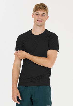Jeune homme portant une chemise noire à manches courtes et un short foncé, souriant, ajustant sa manche droite contre un fond neutre.