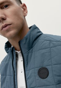 Giacca blu trapuntata con colletto alto, zip completa e un patch circolare nero sul petto. Sotto, è visibile una maglietta polo bianca.