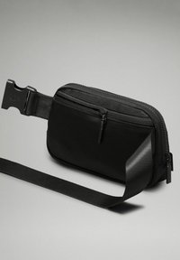 Sac de taille en nylon noir avec un compartiment principal zippé et une sangle détachable. Présente une finition mate et une boucle en plastique robuste.