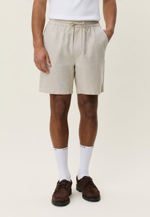 Mand iført beige shorts med snøre, hvide crew-sokker med tekst, og mørkebrune snørebåndssko, står med hænderne i lommerne.