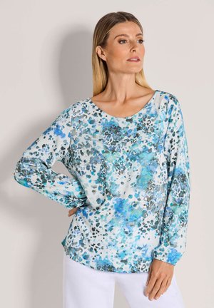 Frau mit glatten blonden Haaren, die eine langärmelige weiße Bluse mit blauen und grauen abstrakten Flecken trägt, kombiniert mit weißen Hosen, steht seitlich.