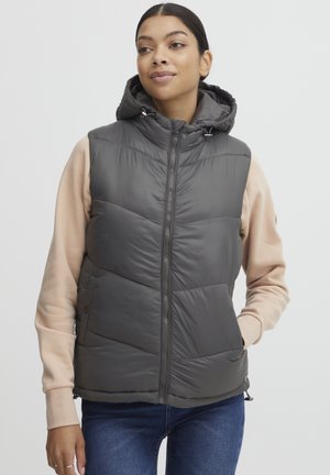 Gilet matelassé gris avec capuche, présentant un zip à l'avant, un tissu texturé, des coutures en chevrons et des poches latérales, porté par-dessus un tee-shirt à manches longues beige.