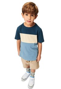 Sininen ja beige väri-blokkaus t-paita lyhyillä hihoilla, yhdistetty beigeihin shortseihin. Lapsella on jalassa valkoiset kengät vaaleansinisillä yksityiskohdilla ja kuvioidut sukat.