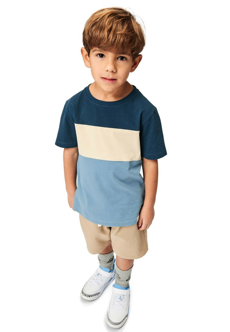 Sininen ja beige väri-blokkaus t-paita lyhyillä hihoilla, yhdistetty beigeihin shortseihin. Lapsella on jalassa valkoiset kengät vaaleansinisillä yksityiskohdilla ja kuvioidut sukat.