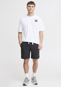 T-shirt blanche à manches courtes avec un graphique noir, associée à un short gris foncé avec poches et un accent logo bleu. Chaussettes blanches et baskets grises.