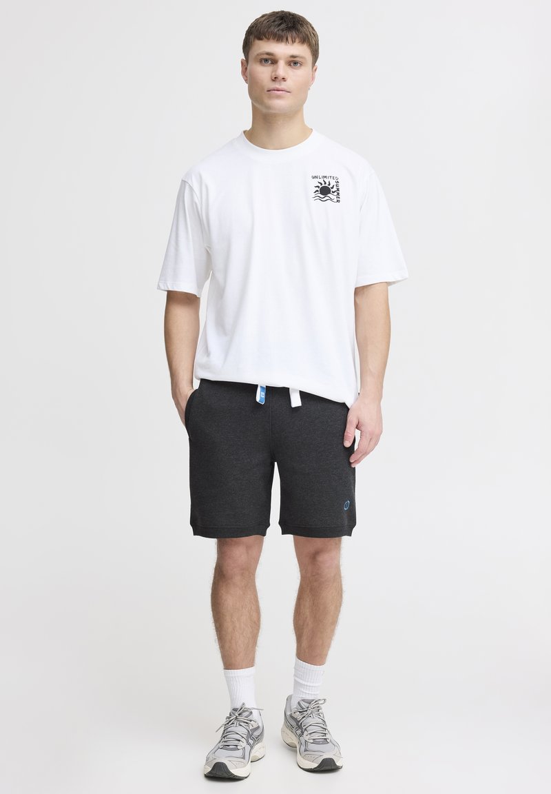 T-shirt blanche à manches courtes avec un graphique noir, associée à un short gris foncé avec poches et un accent logo bleu. Chaussettes blanches et baskets grises.