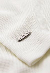 Malá stříbrná visačka s textem "QUINTESSENCE" připevněná k texturované bílé látce s vzorem vaflové tkaniny.