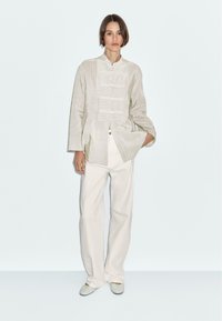 Camicia button-up in lino di colore chiaro con righe verticali, abbinata a pantaloni bianchi larghi. Vestibilità casual, maniche lunghe, design semplice.