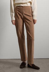 Person wearing brown straight-leg pants, cream sweater, and dark brown slip-on shoes standing on a gray floor against a plain wall.