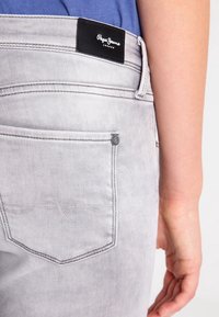 Jeans de mezclilla gris con un parche de cuero negro que presenta el logo "Pepe Jeans London", un bolsillo trasero cosido y sutiles detalles texturizados.