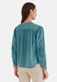 Teehlange Bluse mit langen Ärmeln, gerafftem Rücken, bestickten Details an den Ärmeln, weichem Stoff und geknöpften Manschetten. Schlichtes Rückendesign.