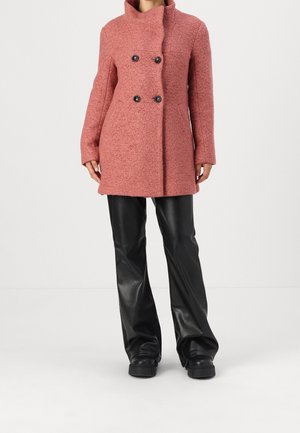 Personne portant un manteau croisé rose poussiéreux texturé avec des boutons noirs, un pantalon en cuir noir évasé et des chaussures compensées noires.