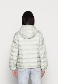 Chaqueta de pluma verde claro con capucha, que presenta un diseño acolchado, puños elásticos y un acabado suave y brillante. Llevada sobre unos jeans remangados.