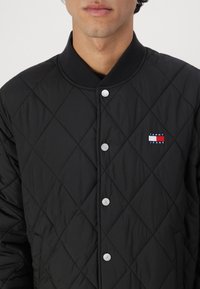Svart quiltet jakke med diamantmønster, med ribbet krage og sølvfargede trykknapper. "TOMMY JEANS"-logo på venstre side.