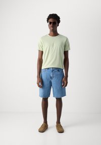 Lys grønn t-skjorte med korte ermer, blå denimshorts, brune slip-on sko og svarte solbriller. Enkel design med løs passform.