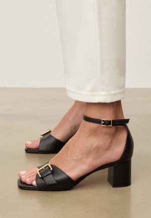 Sandale cu toc bloc negru din piele, cu detaliu de cataramă aurie și curea pentru gleznă, purtate cu pantaloni albi scurtați pe podea bej.