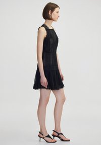Zwarte jurk met kantdetails, aansluitende bodice en uitlopende rok. Doorschijnende stof, ingewikkelde kantafwerking en gecombineerd met zwarte sandalen met bandjes.