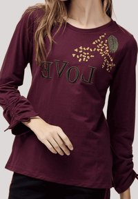 Camiseta de manga larga burdeos con la palabra "AMOR" en negro, que presenta bordados florales en dorado y verde. Mangas fruncidas con lazos ajustables.