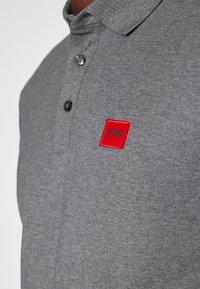 Polo gris hecho de una mezcla de algodón, con un cuello extendido, tres botones y una etiqueta rectangular roja con "HUGO" bordado en negro.