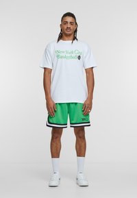 Witte T-shirt met groene tekst "New York City Basketball," gecombineerd met groene mesh shorts met zwarte bies en een decoratief logo.