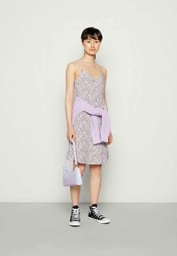 Mulher de pé, vestindo um vestido sem mangas com padrões florais, um suéter lavanda amarrado à cintura, segurando uma mala lavanda e tênis pretos de cano alto.