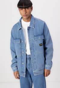 Denim jacket in light blue with a button-front design, two chest pockets, and a black brand patch. Styled with a white t-shirt underneath.