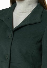 Donkergroene gestructureerde jas met een notch-kraag, twee knopen aan de voorkant en subtiele stiksels. Draagt over een lichte turtleneck.