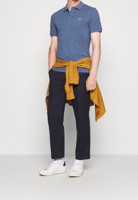 Blå texturerad polo skjorta, marinblå byxor och vita sneakers. Senapsgul skjorta knuten runt midjan. Enkel design med en logotyp på bröstet.