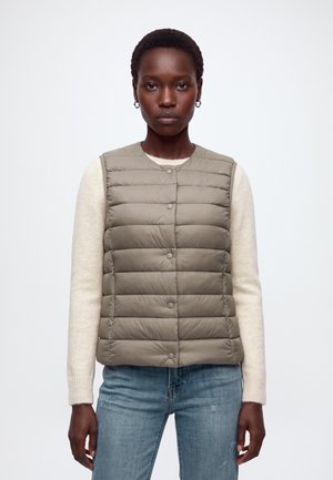 Femme portant un gilet matelassé sans manches beige sur un pull blanc à manches longues et un jean bleu, debout devant un fond blanc uni.