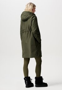 Olivengrøn parka med indsnævret talje, hætte og sidelommer. Bæres over olivengrønne leggings og sorte ankelstøvler med pelsfor. Glat, vandafvisende stof.