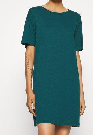 Henkilö, jolla on päällään väljä, lyhythihainen teal-värinen mekko, vasemman kyynärvarren ja käden tatuoinnit näkyvissä, seisoo valkoista taustaa vasten.