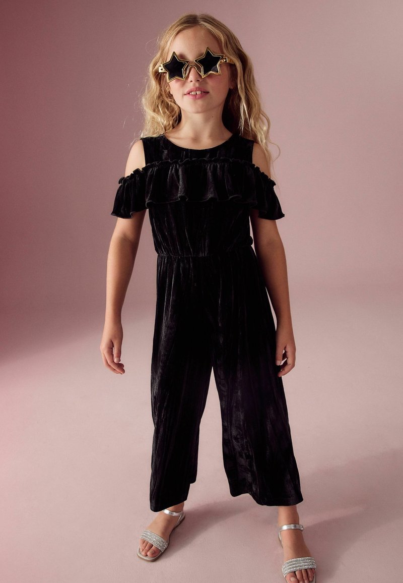 Zwarte fluwelen jumpsuit met schoudervrije ruches, wijde broekspijpen en een elastische tailleband. Gepaard met zilveren sandalen met bandjes en stervormige zonnebril.