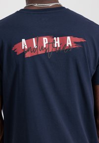 Navyblaue Baumwoll-T-Shirt mit einem roten Pinselstrichdesign, auf dem "ALPHA" in Weiß und "industries" in Grau auf der Rückseite abgebildet ist.