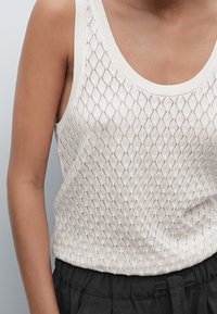 Débardeur blanc en maille avec un motif losange, un large décolleté et des bords côtelés, associé à un pantalon noir texturé.