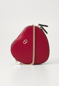 Heart-shaped red leather crossbody bag with gold chain strap, black accents, and logo detail on the front. Smooth texture. Zipper closure.