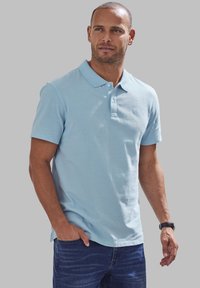 Polo shirt bleu clair à manches courtes en coton, avec un col, trois boutons et un logo discret sur la poitrine. Assorti à un jean foncé.