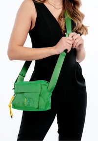 Borsa a spalla verde in rete con chiusura a zip, tracolla regolabile e elemento decorativo giallo. Forma rettangolare compatta con superficie texturizzata.