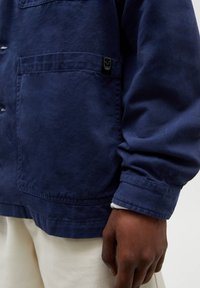 Mörkblå jacka med en slät yta, som har en bröstficka, knäppning och etikettdetalj på framsidan. Vit foder vid ärmsluten.