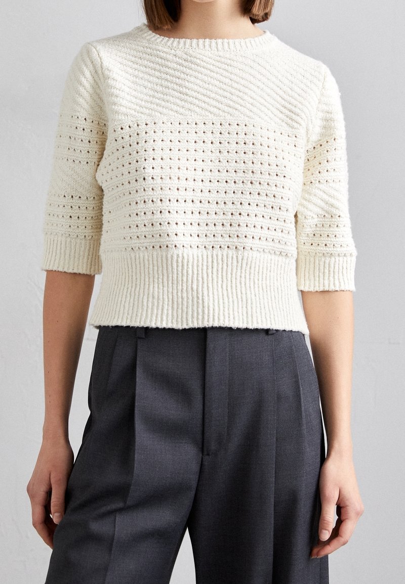 Pull en maille crème à manches courtes, avec un motif texturé et un ourlet côtelé. Associé à un pantalon gris taille haute.