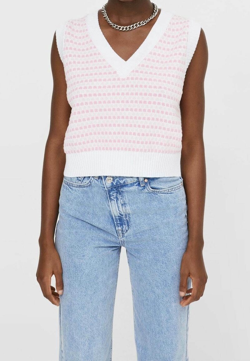 Witte gebreide mouwloze trui met een V-hals, voorzien van een roze stippenpatroon. Gecombineerd met lichtblauwe denim jeans met hoge taille.