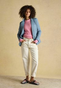 Femeie cu păr creț purtând un pulover roz cu model torsadat, sacou albastru, pantaloni crem și pantofi slip-on colorați, stând cu mâinile în buzunare.