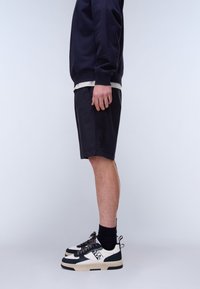 Persoon staand in zwarte shorts, marineblauwe trui, zwarte sokken en zwart-witte sneakers tegen een effen lichte achtergrond.