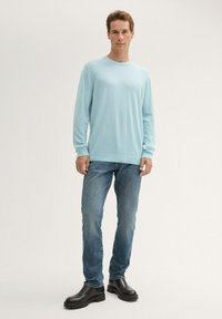 Jasnoniebieska koszula z długim rękawem z teksturowanej tkaniny, w zestawieniu z wyblakłymi niebieskimi dżinsami i czarnymi butami na grubym obcasie, stojąca na neutralnym tle.