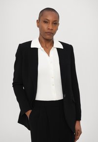Svart blazer med strukturerte skuldre, brukt over en tettsittende hvit ribbestrikket topp med krage. Har knapper og en skreddersydd passform. Enkel bakgrunn.
