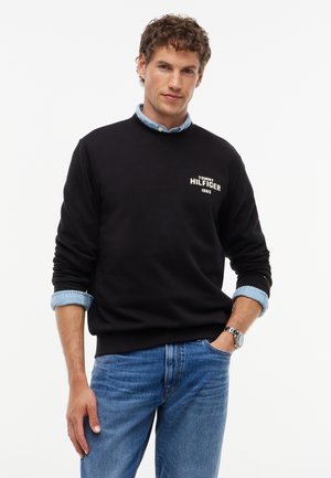 Homme portant un sweat-shirt noir avec le texte "Tommy Hilfiger 1985", manches de chemise bleu clair retroussées, jean bleu et une montre-bracelet, debout avec une main dans la poche.