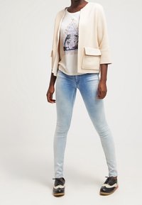 Chaqueta beige clara con mangas cortas y un bolsillo frontal, combinada con una camiseta gráfica blanca y unos vaqueros ajustados de color azul claro. Zapatillas en blanco y negro.