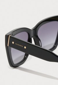Ochelari de soare de tip cat-eye, negri, cu lentile gradient de culoare purpurie și marcaj de aur "ALLSAINTS" pe bradă, pe un fundal alb.